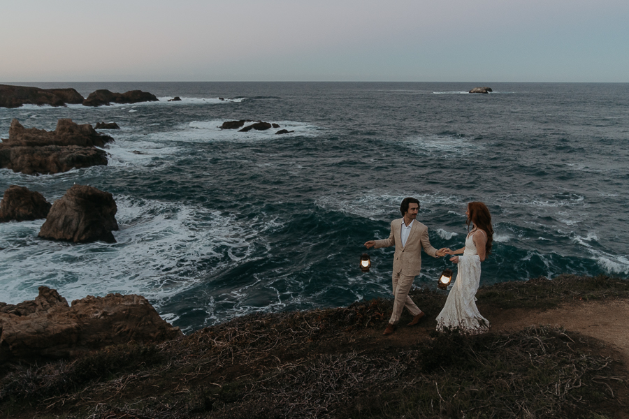 Bride and groom wearing wedding attire walking down a trail hand in hand holding lanterns by the ocean 