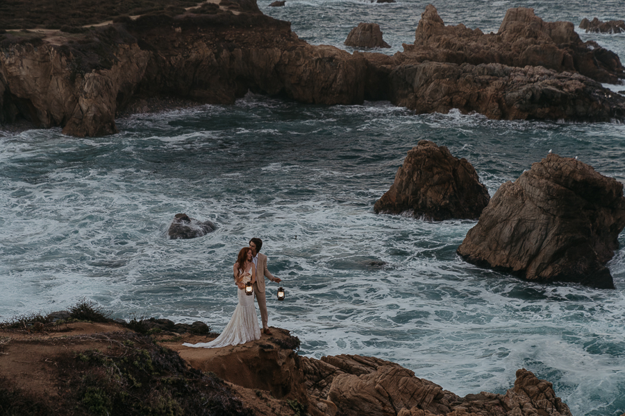 Bride and groom holding each other at sunrise in Big Sur on a trail overlooking the ocean by light of lantern