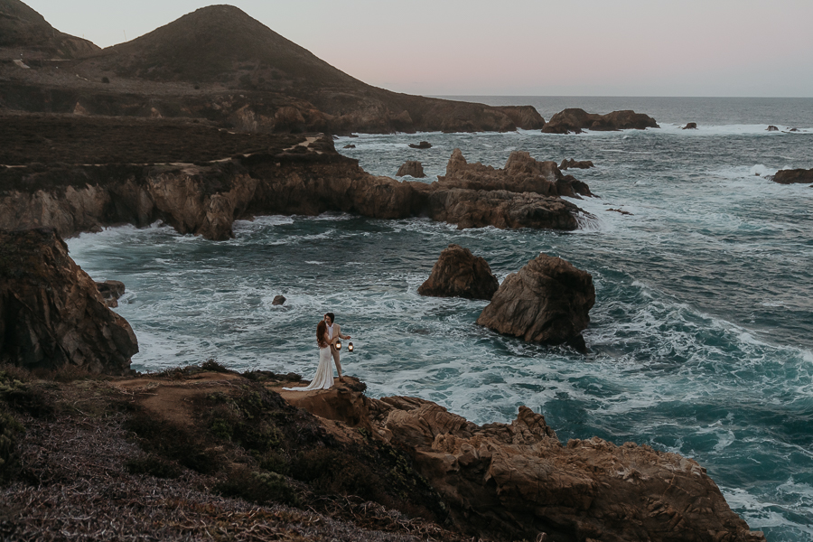 Bride and groom wearing wedding attire holding lanterns overlooking the ocean on a bluff trail in Big Sur at sunrise taken by Big Sur elopement photographer Kasey Mantiply