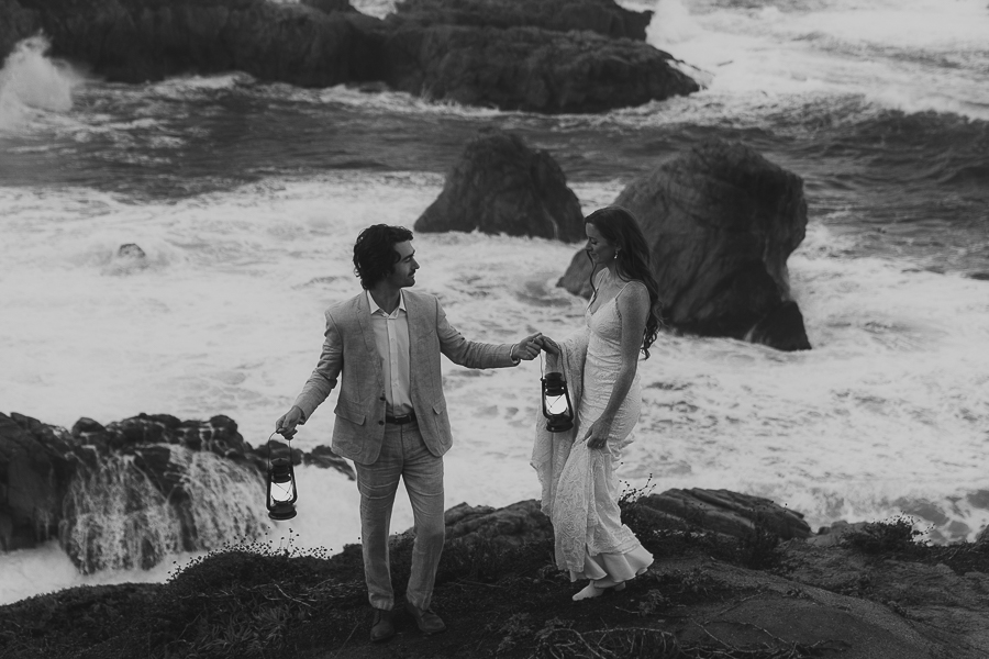 Black and white photo of bride and groom in wedding attire walking on a bluff trail in Big Sur while holding lanterns