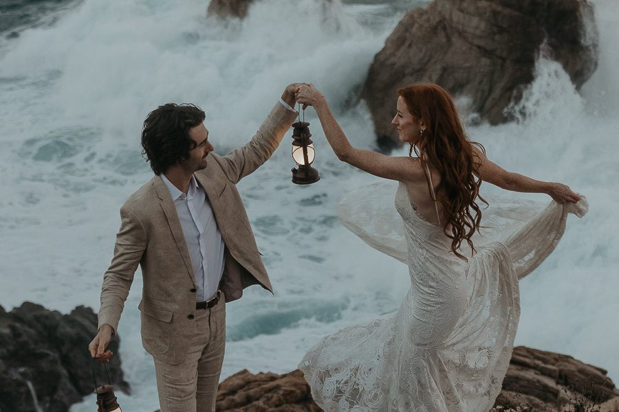 Bride wearing lace gown and groom wearing tan suit dance on a bluff above the ocean groom twirls bride around while holding a lantern and bride's gown flows while twirling