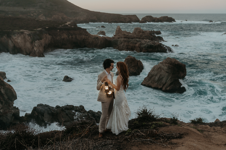 Bride in lace gown and groom in tan suit looking at each other holding lanterns illuminating their faces on a cliff above the ocean in Big Sur at sunrise