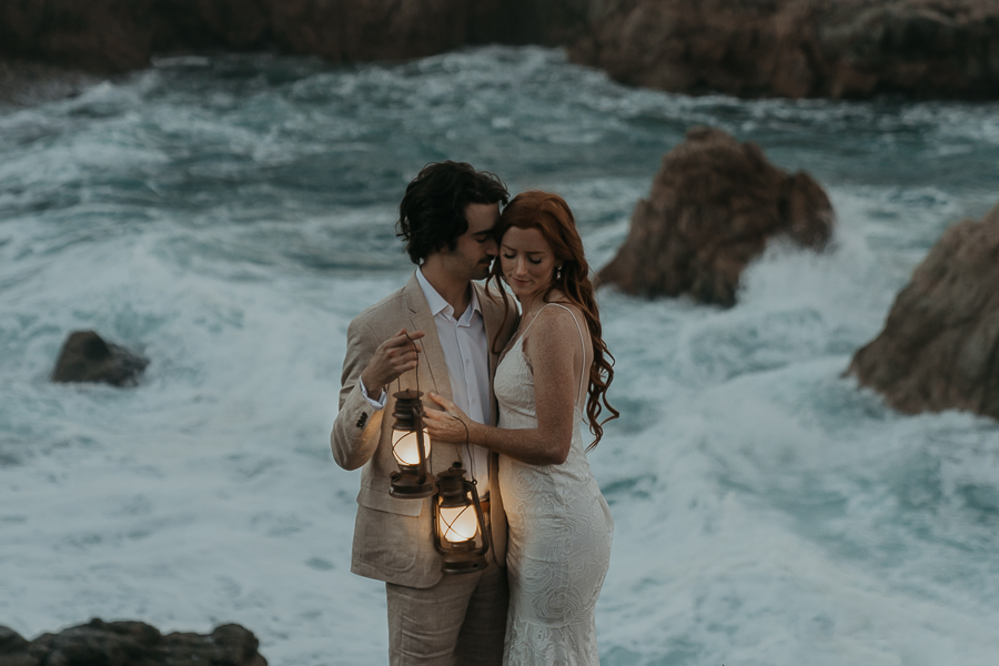 Bride and groom wearing wedding attire groom nuzzles brides face bride smiles looking down while they both hold lanterns illuminating them at sunrise in Big Sur