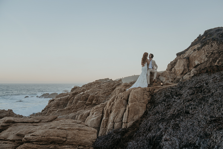 Bride and groom standing on rocks above the ocean groom is sitting down while bride stands and holds his face 