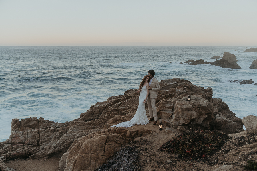 Bride and groom stand on rocks above the ocean with lanterns on the rocks by their feet