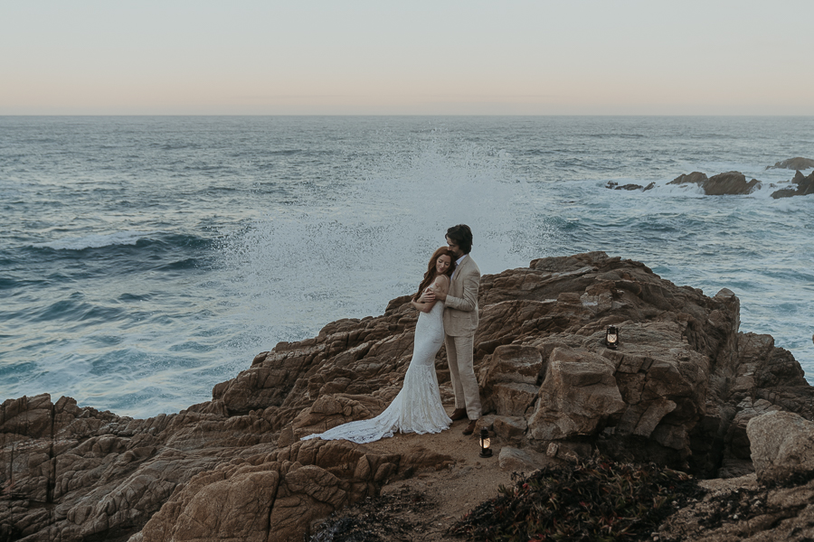 Groom holds bride while bride faces away and turns head toward groom while wave splashes up on the rocks behind them in Big Sur