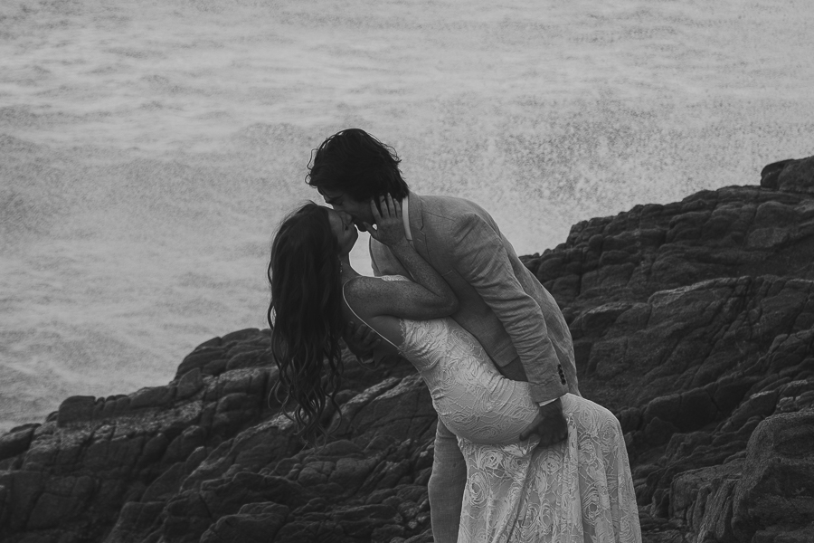 black and white photo of bride and groom in a dip kiss on rocks in front of the ocean with wave splashing up behind them
