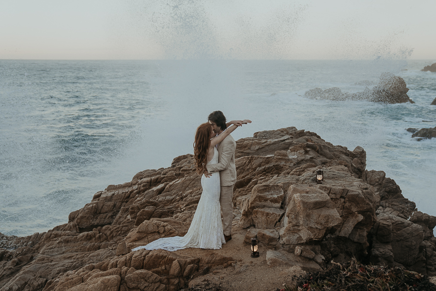 Bride wearing lace gown and groom wearing tan suit kiss on rocks by the ocean while wave splashes up behind them