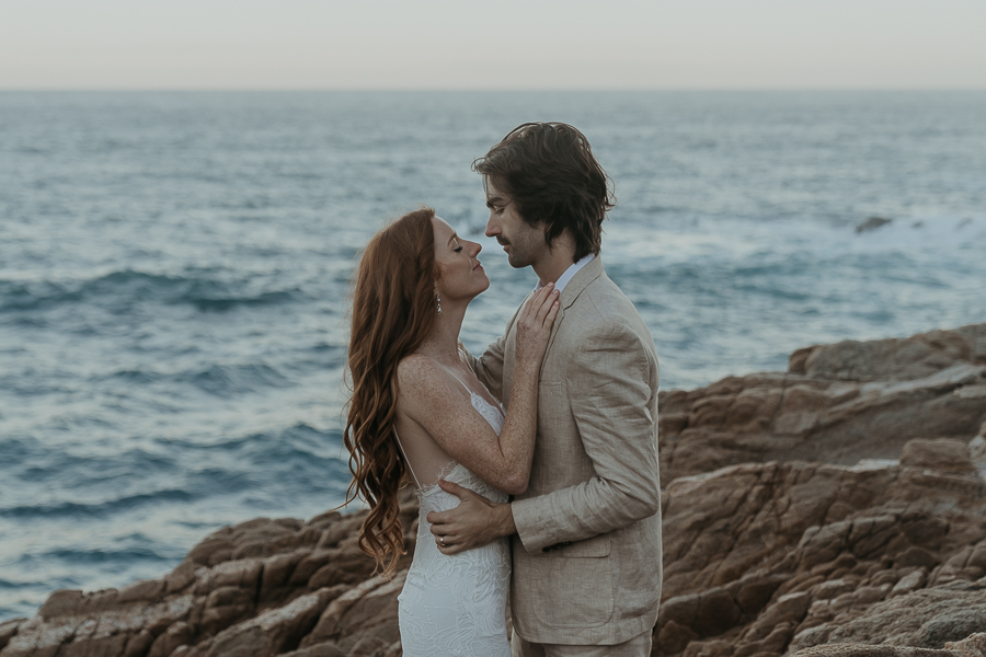 Bride and groom looking at each other standing on rocks in front of the ocean