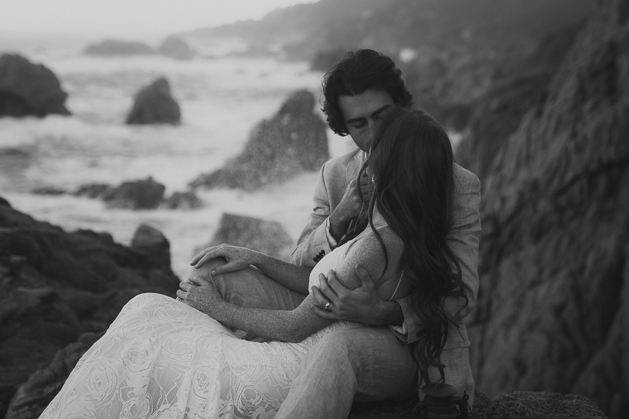 Black and white photo of bride and groom kissing while sitting on rocks by the ocean
