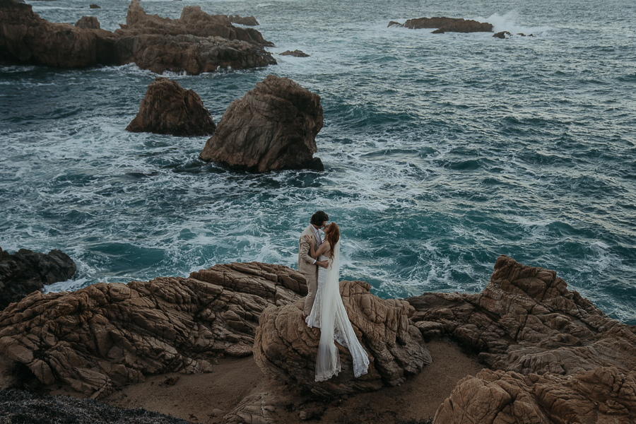 Bride and groom standing on rock in front of the ocean kissing while brides veil flows down the rock