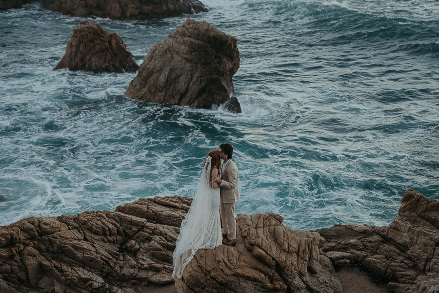 Bride wearing lace gown and long veil kisses groom wearing tan suit standing on rock in front of the ocean