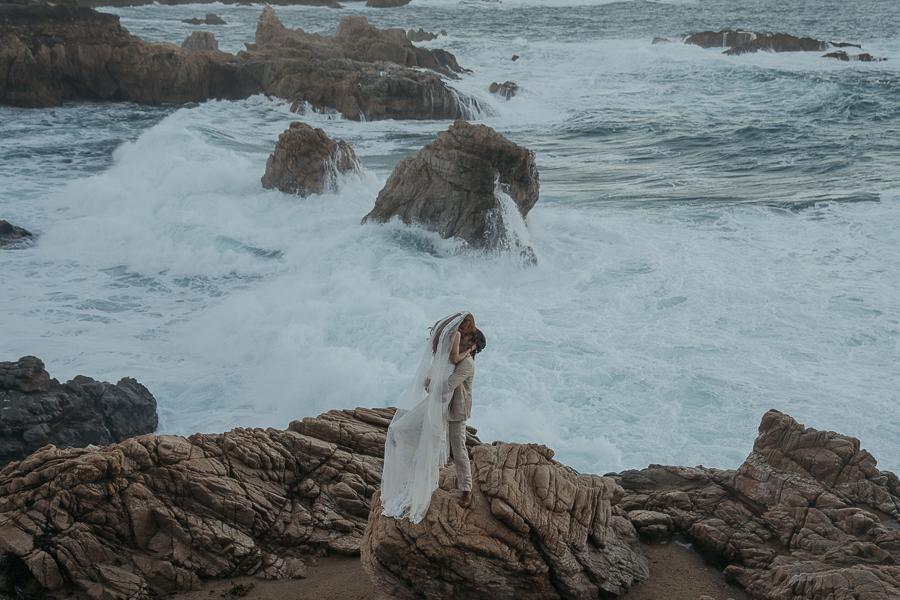 Groom wearing tan suit lifts bride off the ground while kissing standing on rock in front of the ocean after eloping in Big Sur