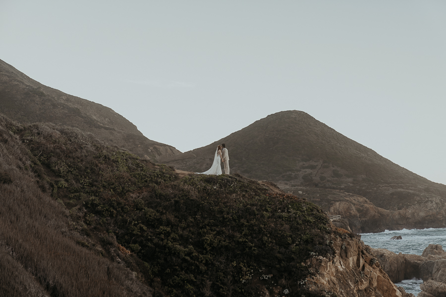 Bride and groom hold both hands and lean in for a kiss standing on a cliff above the ocean in Big Sur with hill in background