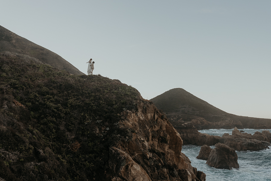 Groom lifts bride into air while bride holds out her arms standing on cliff far away overlooking the ocean with hills in the background