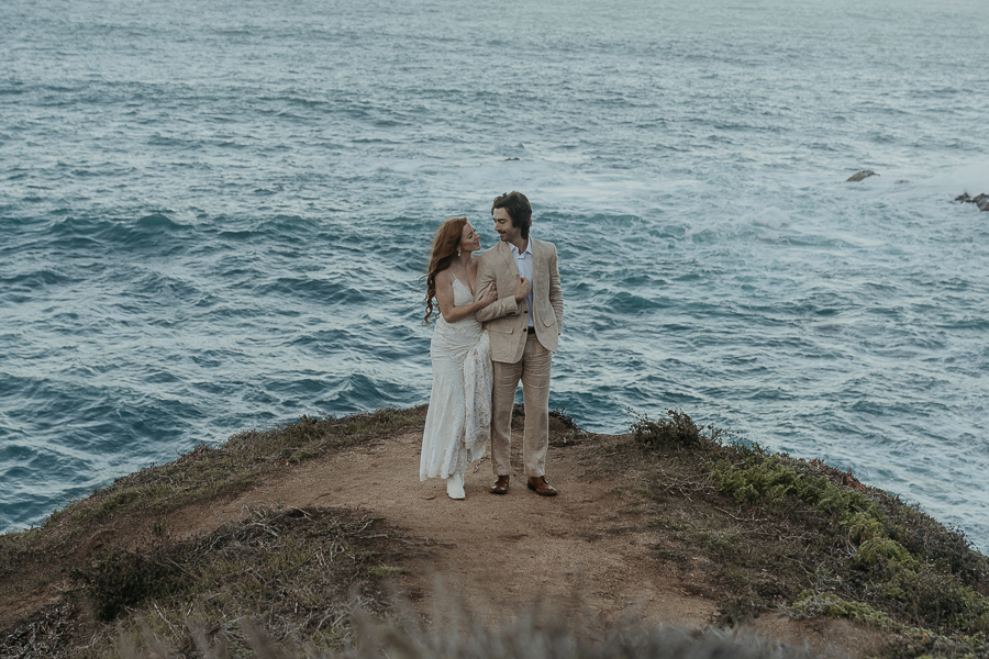 Bride wearing lace gown and white boots holds onto grooms arm while walking up trail on bluff above the ocean in Big Sur