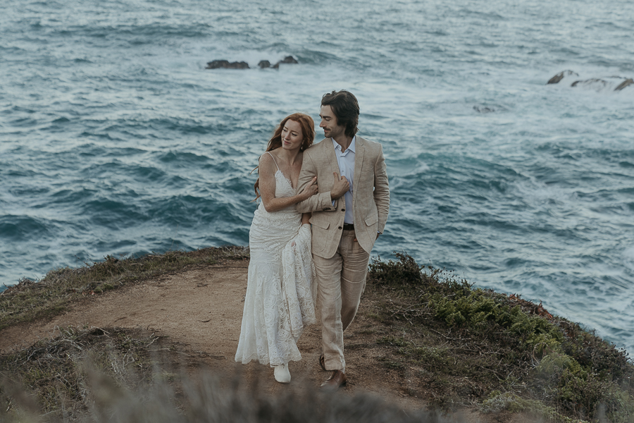Bride wearing lace gown and white boots holds grooms arm while walking on bluff trail above the ocean 
