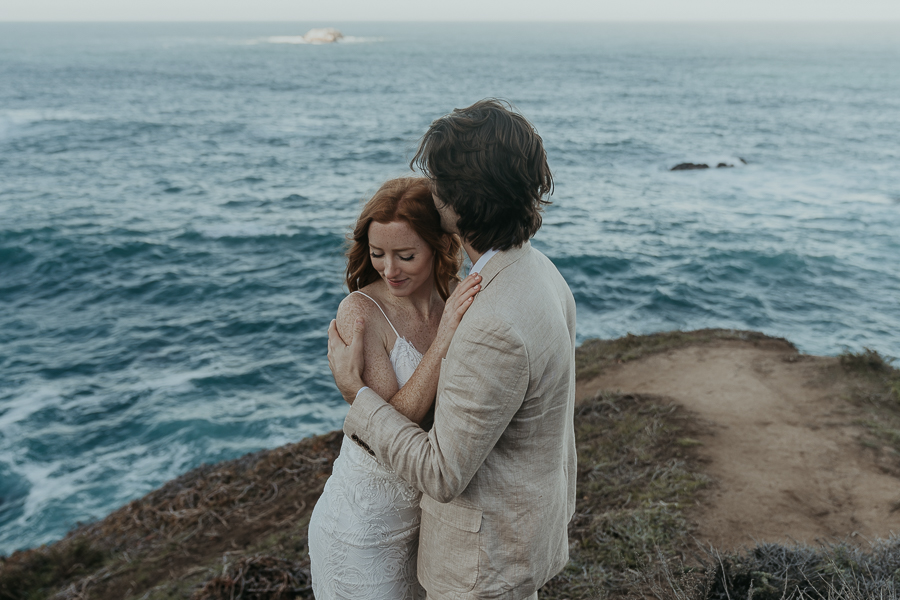 Groom wearing tan suit kisses the side of brides head standing on a bluff trail above the ocean 
