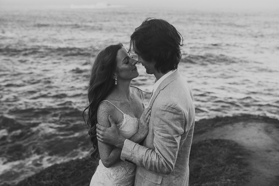 Close up black and white photo of bride and groom touching noses standing on bluff above the ocean