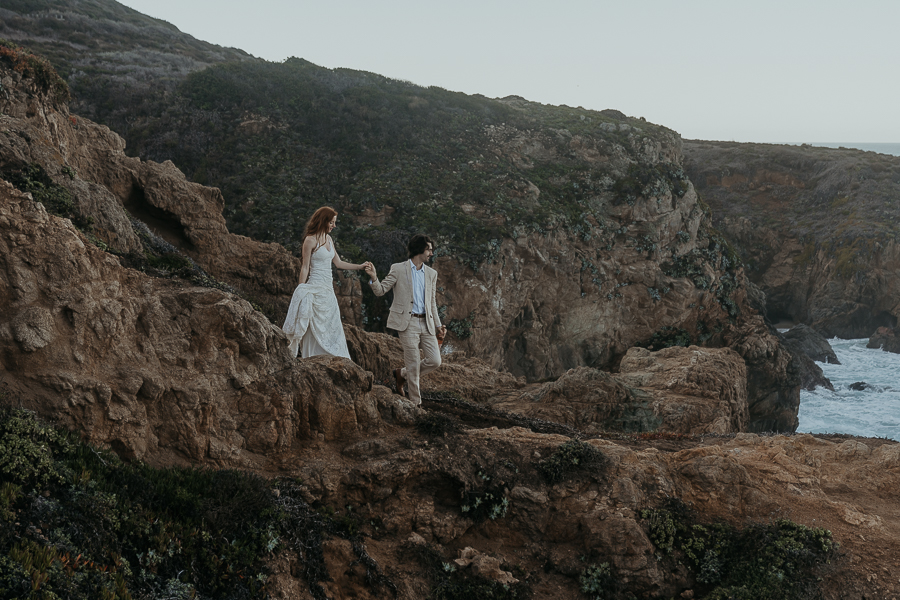 Groom wearing tan suit leads bride wearing lace gown down trail on coastal bluffs in Big Sur