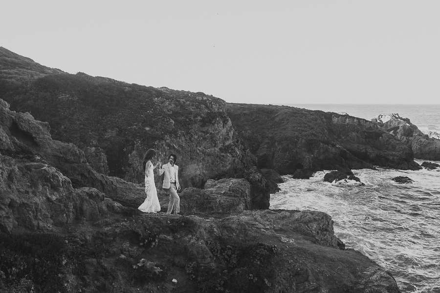 Black and white photo of bride and groom wearing wedding attire walking down rocky trail towards the ocean in Big Sur