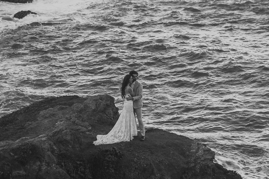 Black and white photo of bride and groom hugging standing on coastal bluff above the ocean