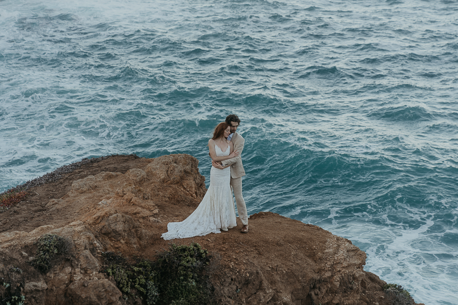 Bride and groom holding each other standing on cliff above the blue ocean in Big Sur