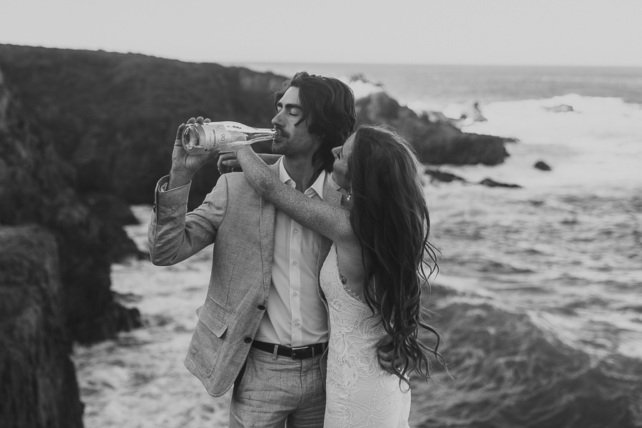 Black and white photo of bride and groom hugging while groom drinks from champagne bottle with coastal bluffs and ocean in the background
