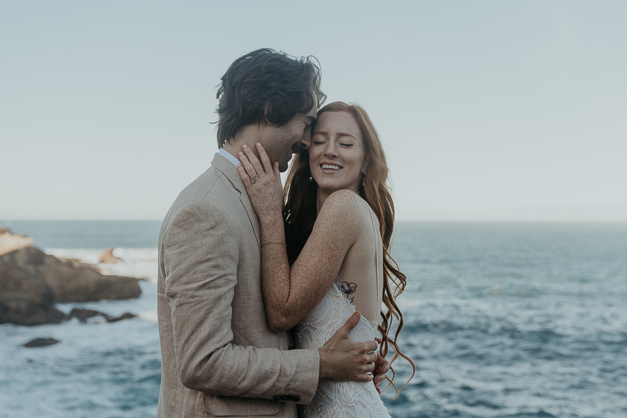 Close up photo of bride smiling while groom leans against her face smiling in front of ocean