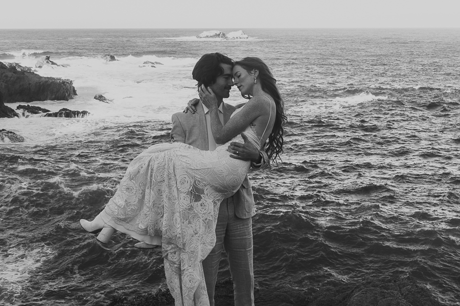Black and white photo of groom picking up bride and holding her while smiling at her and bride holds grooms face with ocean in the background