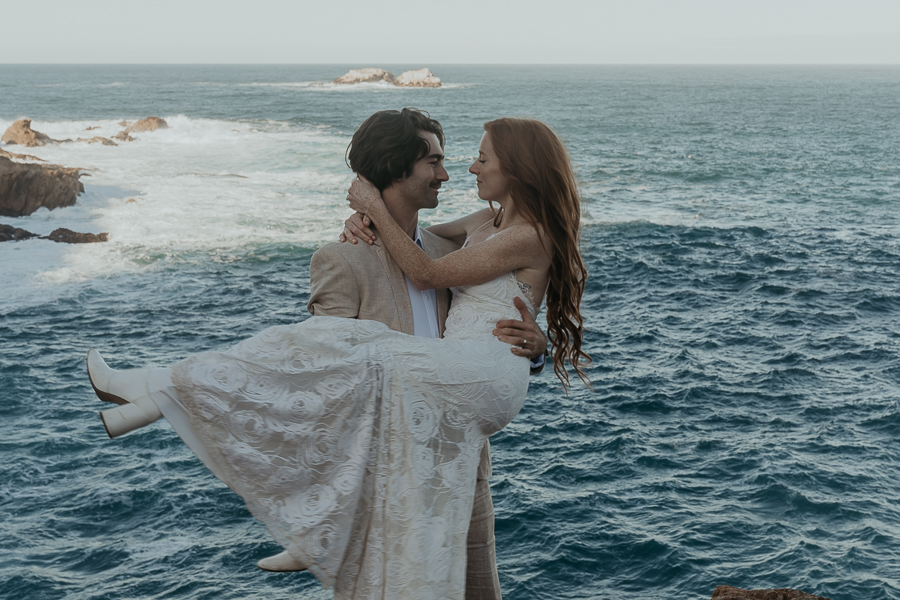 Close up photo of groom wearing tan suit picking up and holding bride wearing lace gown and white boots with arms wrapped around grooms neck with ocean in the background