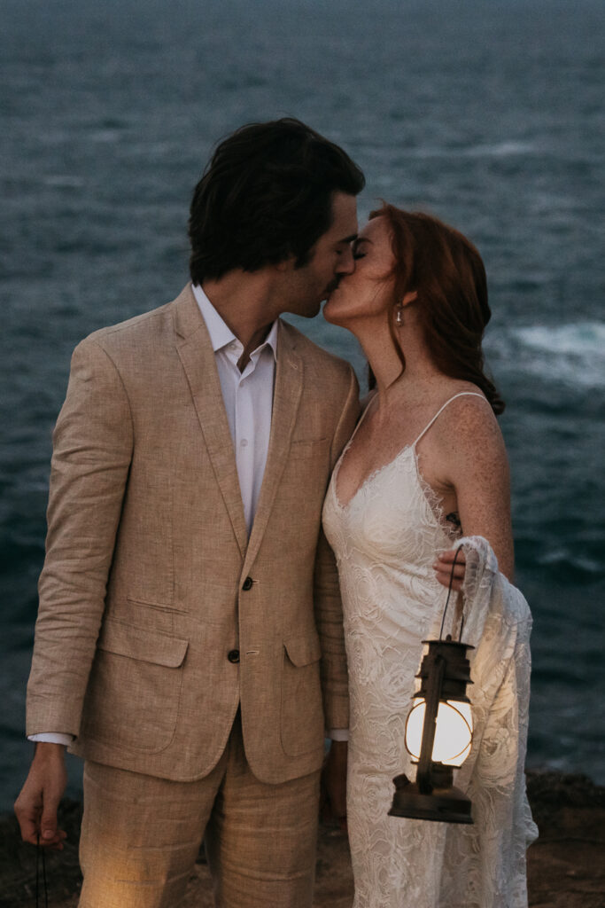 Bride and groom kissing holding lanterns in front of ocean