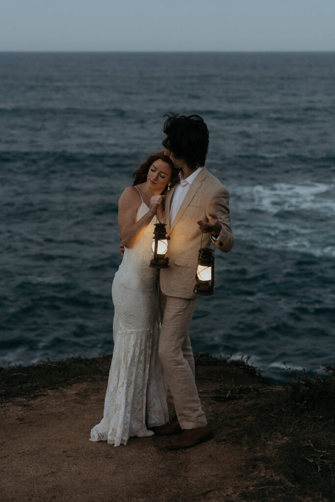 Bride leaning head on groom's chest holding lanterns in front of ocean