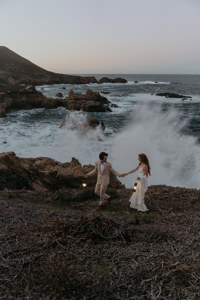 Bride and groom wearing wedding attire holding lanterns walking down a trail on a cliff over the ocean in Big Sur at sunrise