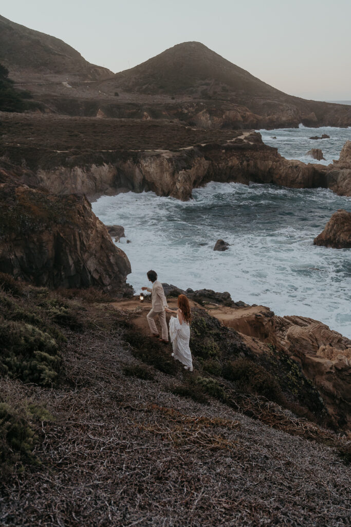 Groom leading bride down a hiking trail by lantern on the Big Sur coast