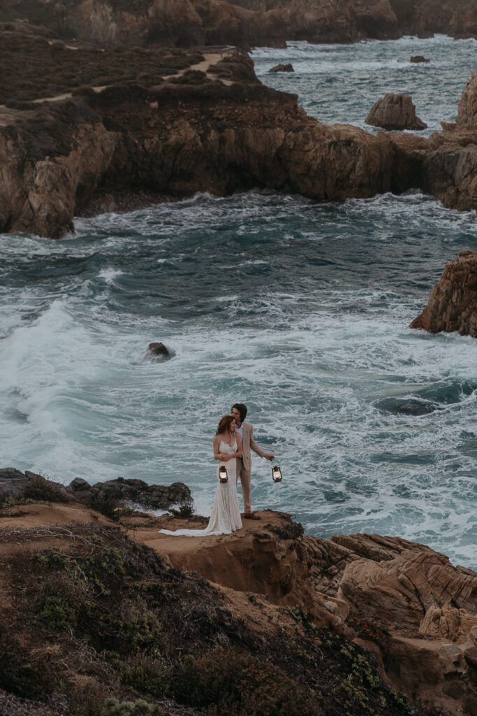 Groom holding brides waist while holding lanterns overlooking the ocean in Big Sur