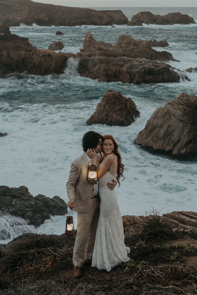 Groom wearing a tan suit and bride wearing a lace gown holding each other while smiling and bride looks away holding lanterns on a cliff above the ocean in Big Sur