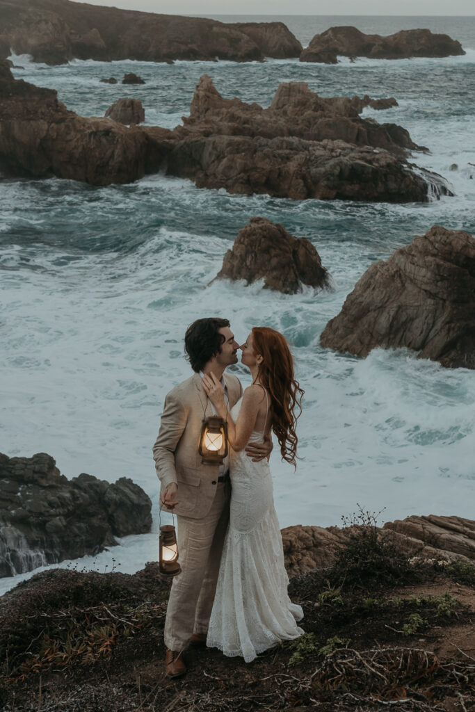 Bride wearing a lace wedding dress and groom in a tan suit holding each other and touching noses while holding lanterns on a bluff overlooking the ocean in Big Sur