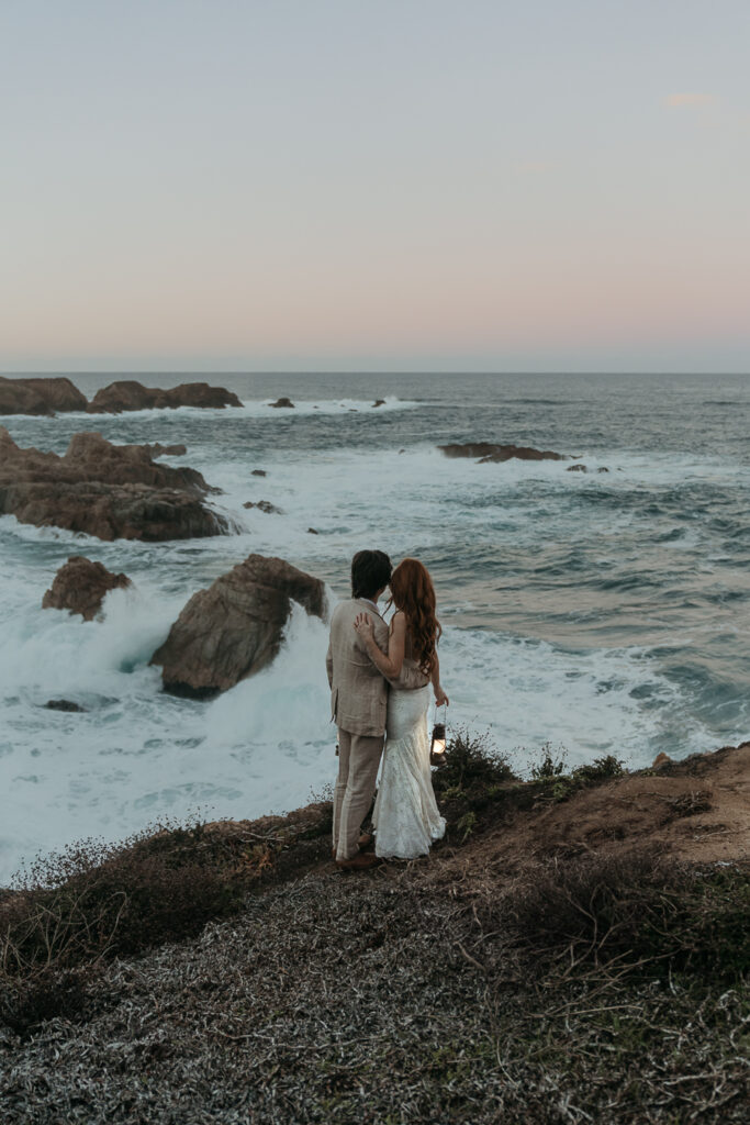 Bride and groom in wedding attire holding each other's waist looking away from the camera over the ocean in Big Sur