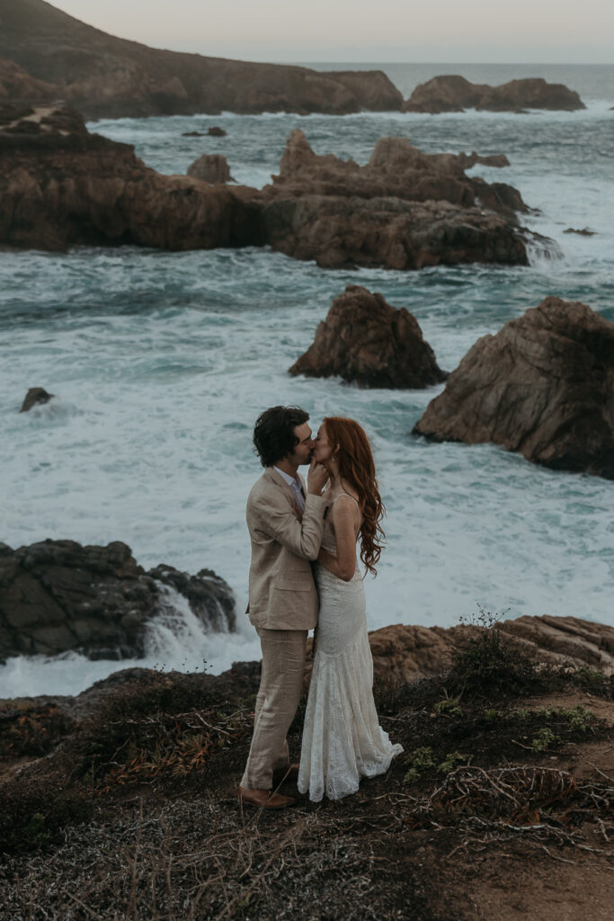 Bride wearing a lace gown and groom wearing a tan suit kissing on a bluff overlooking the ocean in Big Sur