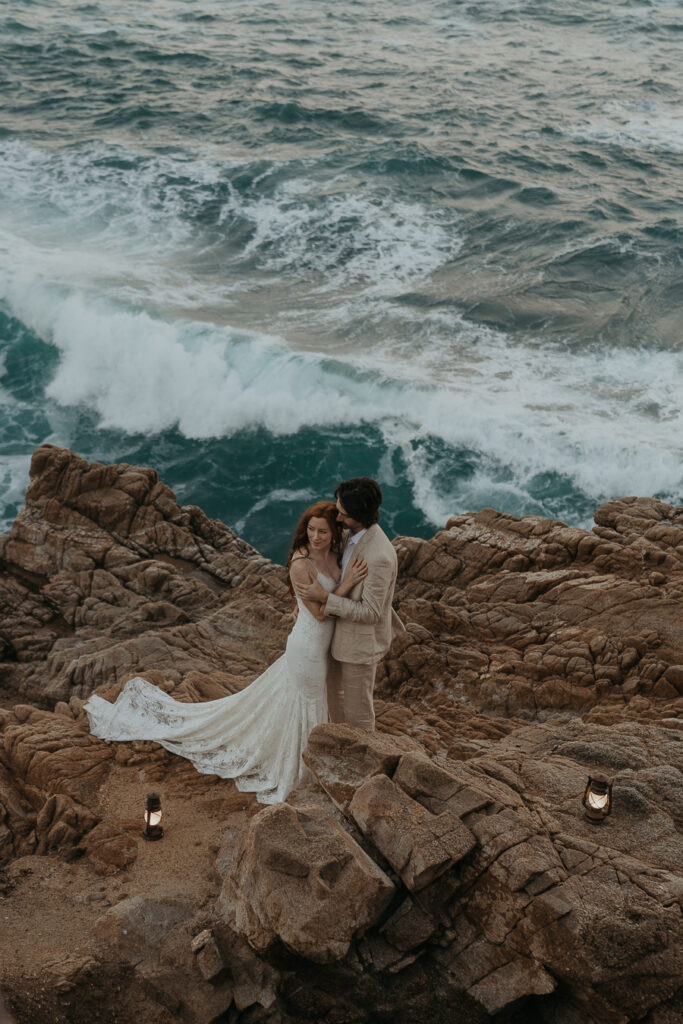 Bride and groom holding each other on rocks above the ocean with lanterns on either side of them placed on the rocks
