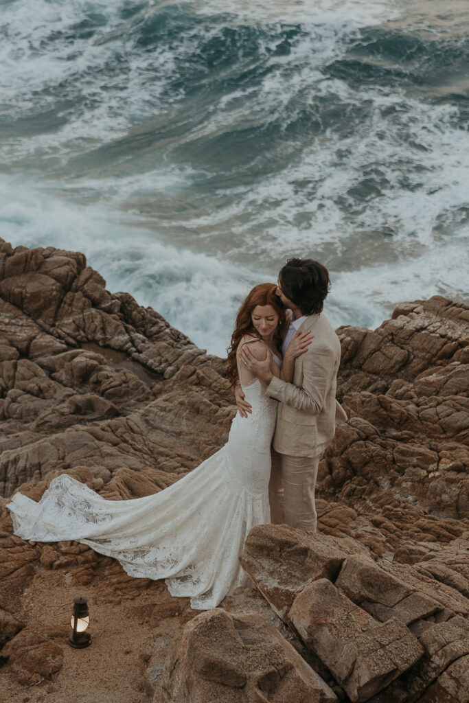 Bride in lace dress hugs groom in tan suit on rocks in front of the ocean with lantern on the ground next to them