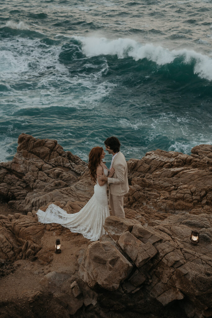 Bride and groom wearing wedding attire looking at each other facing each other with ocean in the background and lanterns on the rocks by their feet