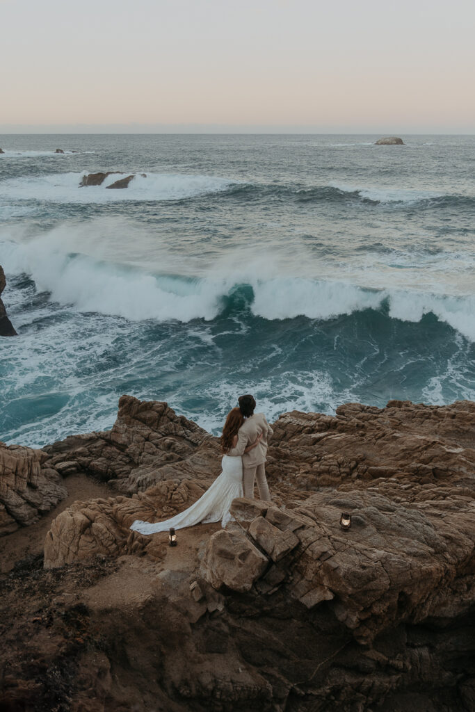 Bride wearing lace gown and groom wearing tan suit stand on rocks above the ocean holding each other facing away from the camera looking at the ocean and pink sunrise sky with lanterns on the rocks behind them. 