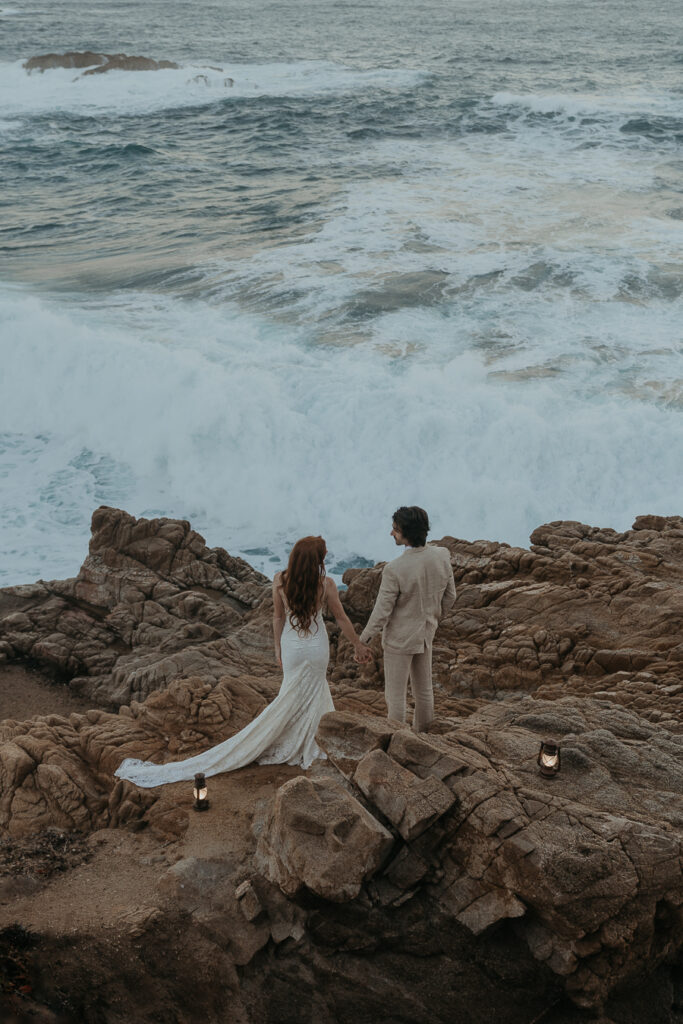 Bride and groom looking at each other holding hands in front of the ocean with lanterns placed on the rocks behind them