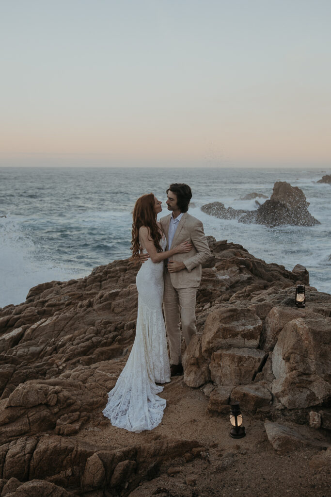 Bride and groom facing each other looking at each other standing on rocks above the ocean in Big Sur with lanterns on the rocks by their feet