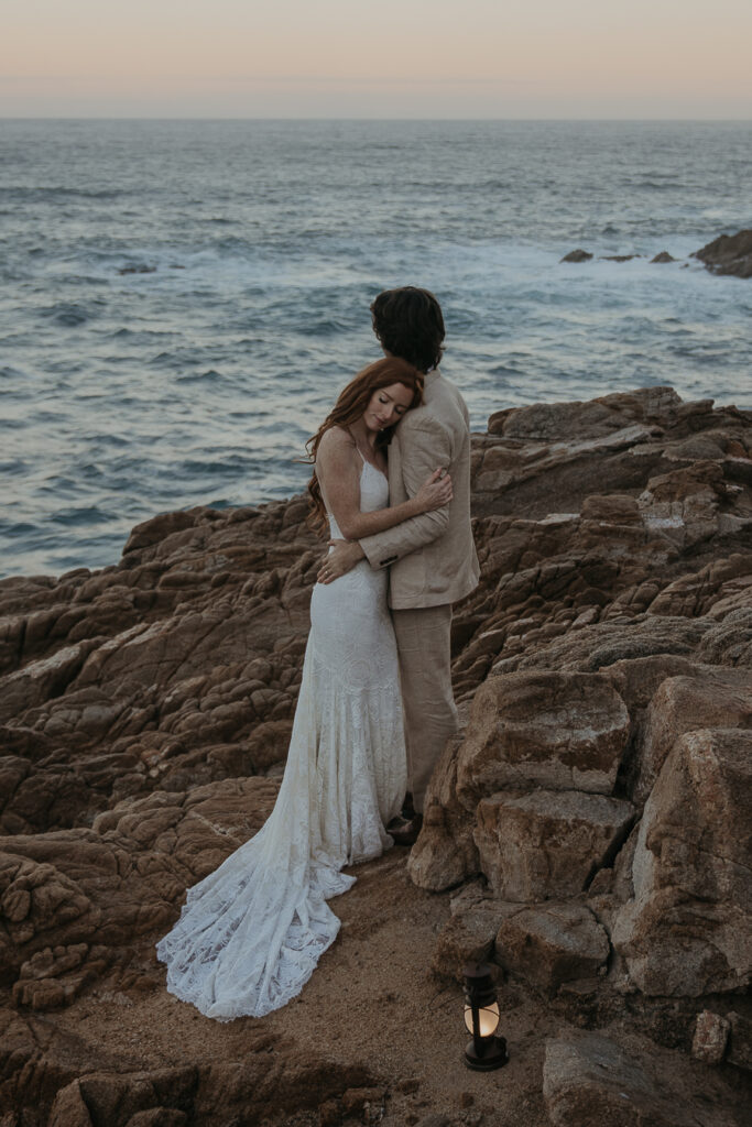 Groom hugs bride while bride rests her head on grooms shoulder in front of the ocean at sunrise in Big Sur