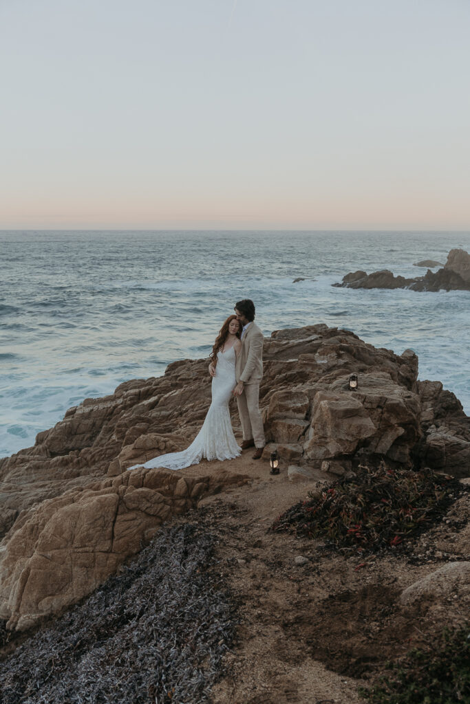 bride and groom stand on rocks in front of the ocean with lanterns by their feet