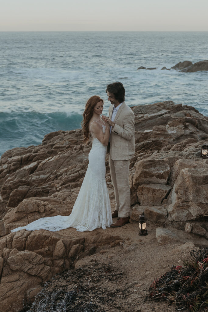 Bride and groom in wedding attire facing each other holding hands while looking at their hands standing on rocks above the ocean in Big Sur at sunrise with lanterns on the rocks by their feet