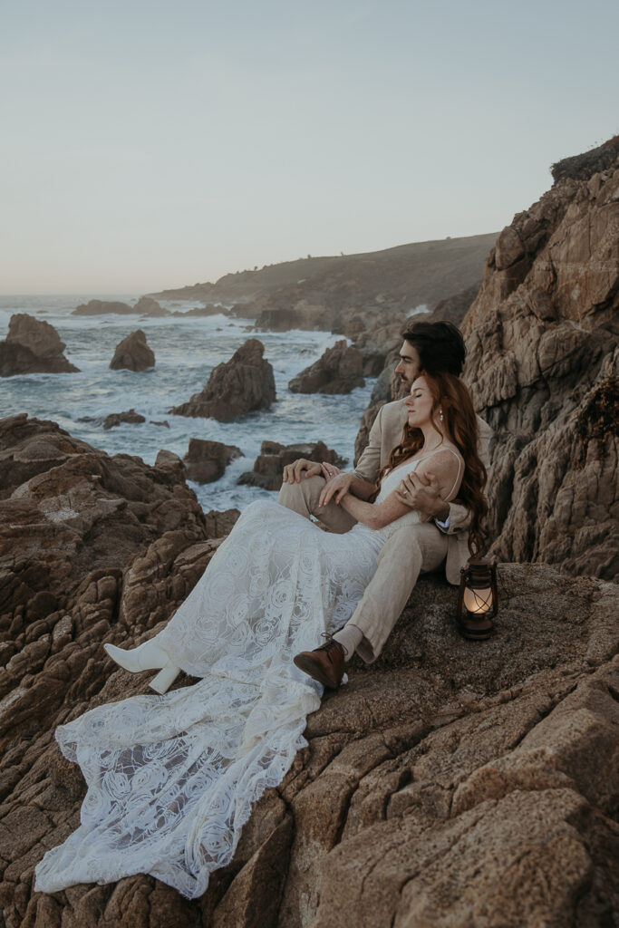 bride wearing lace dress and white boots sits in grooms lap on rocks overlooking the ocean